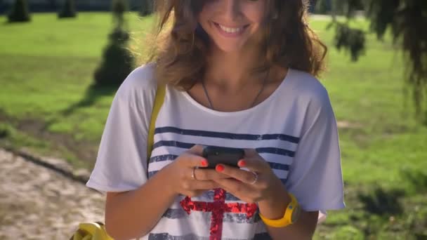 Porträtt av en ung leende kaukasiska flicka stående i parken och använder en smartphone universitet i bakgrunden — Stockvideo