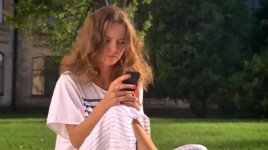 Bir smartphone kullanarak kaydırma, yazarak, üniversite içinde belgili tanımlık geçmiş çim üzerinde parkta oturan genç beyaz esmer