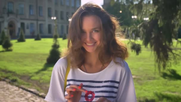 Porträt eines jungen lächelnden kaukasischen Mädchens, das im Park steht und ein Smartphone benutzt und im Hintergrund die Universität scrollt — Stockvideo