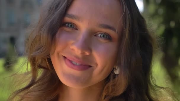 Porträt einer jungen lächelnden kaukasischen Brünette im Park, im Hintergrund die Universität — Stockvideo