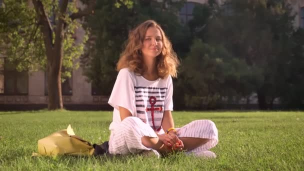 Junge lächelnde kaukasische Brünette sitzt im Park auf Gras, blickt in die Kamera, lacht, Universität im Hintergrund — Stockvideo