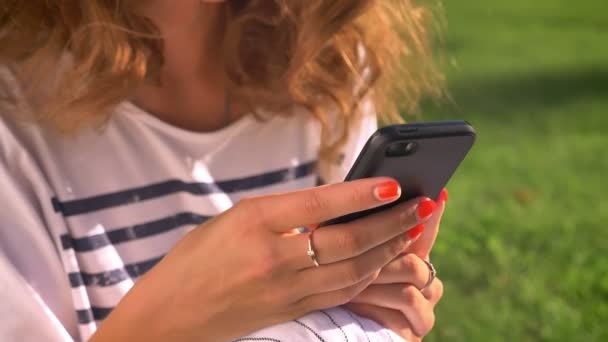 Unga kaukasiska brunett sitter i parken på gräset, som använder en smartphone, rullning, maskinskrivning, universitet i bakgrunden — Stockvideo
