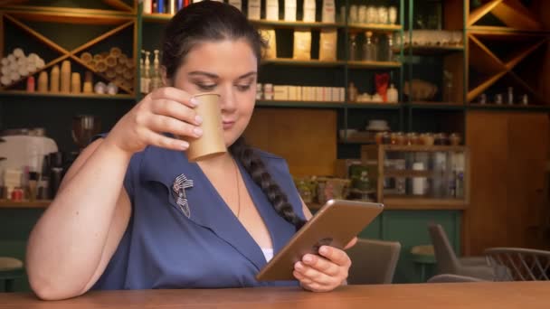 Grande caucásico sonriente chica deslizando su planshet y beber café en la mesa — Vídeos de Stock