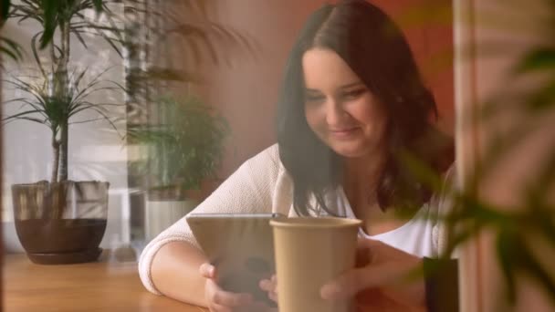 Добре нагодована смішна біла дівчина дивиться на планшет і має каву поруч з вікном — стокове відео