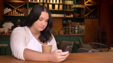 İçecek fincan ile kafede otururken tutan smartphone swiping yusyuvarlak beyaz kadın
