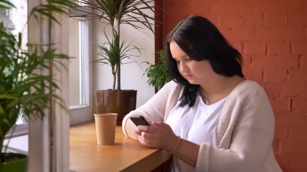 Пухнаста біла дівчина прокручує і друкує на смартфоні на столі в кафе з чашкою напоїв — стокове відео