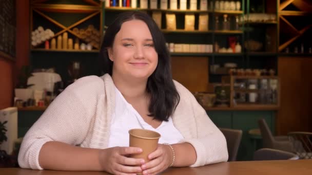 Fubsy ragazza caucasica guardando la fotocamera e sorridente tenendo grande tazza di caffè al coperto — Video Stock