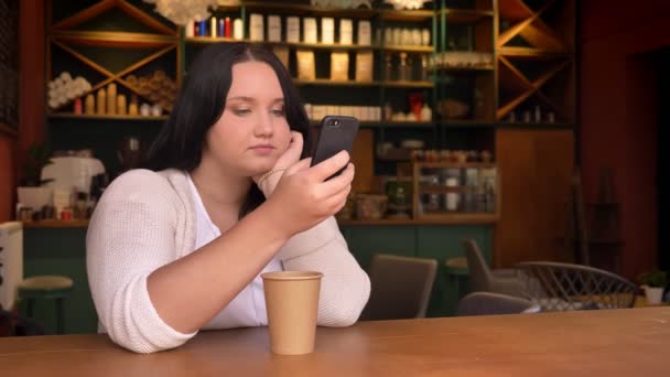 Schwere kaukasische Frau wischt ihr Handy und sitzt im Café mit großen Pappbecher — Stockvideo