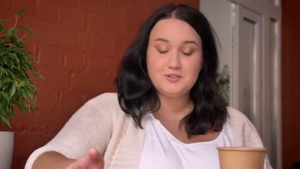 Overweighted caucasian talkative female holding cup of coffe and energically waving her hand in cafe — Stock Video