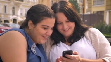 İki büyük beyaz kadın açık bankta otururken swiping smartphone gülümseyen