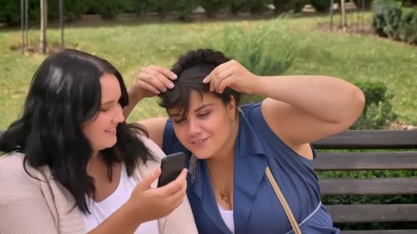 Dos mujeres caucásicas pesadas que se divierten y endurecen el cabello mientras toman selfie relajarse en el banco en el parque — Vídeos de Stock