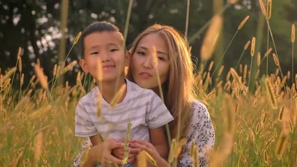 Smiling asian mother with her little son sitting and hugging in grass on what field landscape — Stock Video
