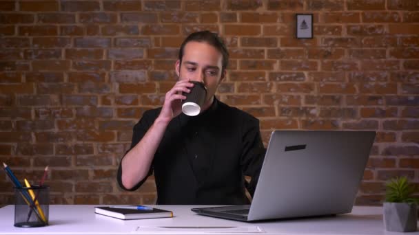 有吸引力的年轻白种男性喝咖啡和看计算机在他的工作地点在红色演播室里面 — 图库视频影像