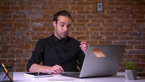 Розчарований білий чоловік в чорному костюмі покриває обличчя руками, дивлячись на ноутбук і сидячи біля цегляної стіни — стокове відео