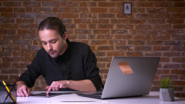 Atractivo caucásico hombre adulto deslizando su tableta y mirando a la computadora mientras está sentado en la oficina de ladrillo en traje negro — Vídeos de Stock