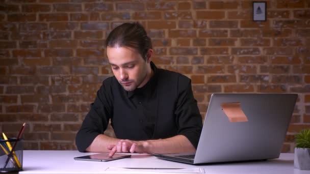 Lindo tipo caucásico desplazando su tableta mientras está sentado en su lugar de trabajo con pared de ladrillo detrás — Vídeos de Stock