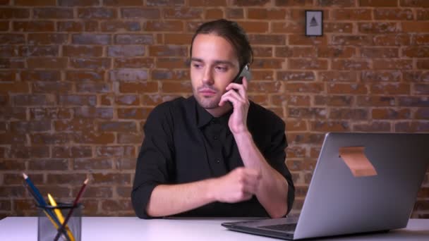 Pohledný mladý muž se soustředěním drží telefon a čeká na odpověď při pohledu na počítači, zatímco sedí v kanceláři cihla — Stock video