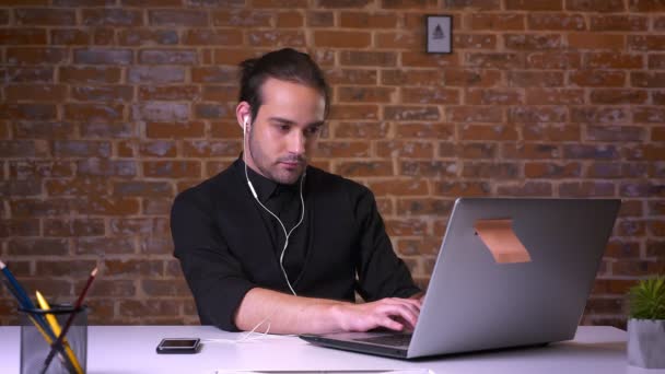 Détendu mignon caucasien gars dans les écouteurs écouter de la musique et dactylographier sur son ordinateur portable à côté de mur de briques en studio — Video