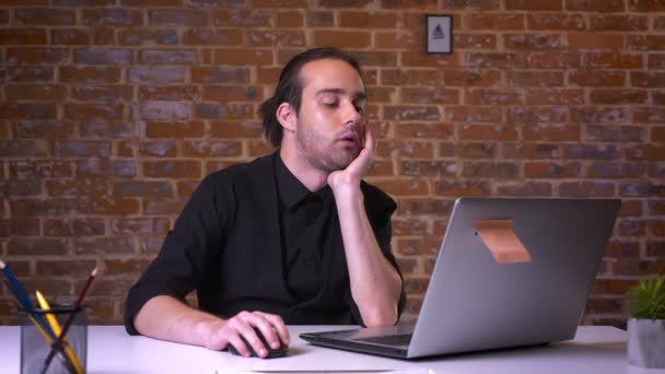 Bored caucasian businessman yawning while looking at laptop and sitting at desk relaxed with brick background — Stock Video