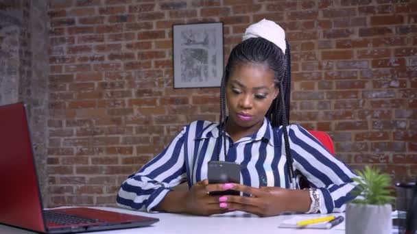 Enjouable Afrikaanse zakenvrouw toont groen scherm en glimlacht gelukkig en ontspannen bij de balie naast de bakstenen muur — Stockvideo