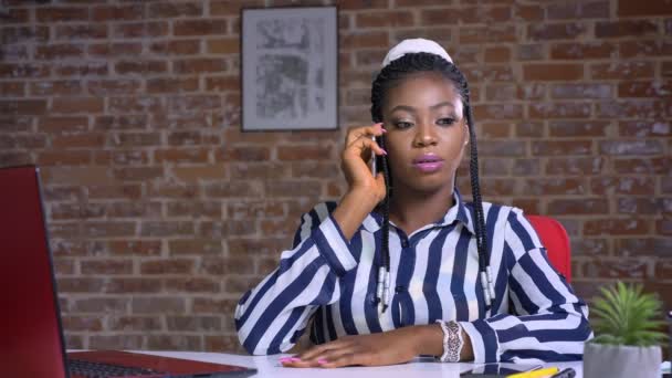 Serious african girl hanging over phone and talking while sitting at the table on brick background — Stock Video