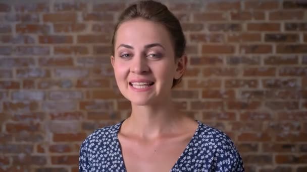 Baumelndes selbstbewusstes Lächeln einer jungen kaukasischen Frau, die still steht und in die Kamera mit ziegelroter Wand hinter sich blickt — Stockvideo