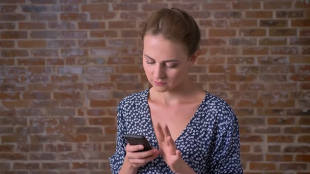Söt kaukasiska kvinna att svepa hennes telefon lätt och tittar på kameran med förtroende på tegel bakgrund — Stockvideo