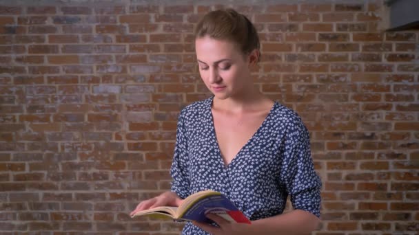 Happy calm caucasian brunette fwoman is standing straight, reading book and looking at camera relaxed on brick wall — Stock Video
