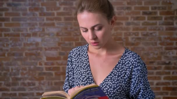 Концентрована брюнетка біла жінка тримає книгу, читає і дивиться з посмішкою на камеру на цегляному фоні — стокове відео