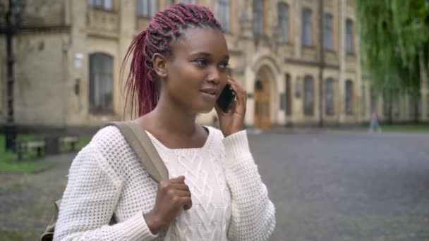 Молодая очаровательная африканская студентка разговаривает по телефону и держит рюкзак, стоя в парке рядом с университетом — стоковое видео