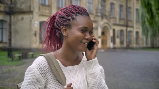 Joven africana hermosa estudiante hablando por teléfono y riendo, la celebración de la mochila y de pie en el parque cerca de la universidad, feliz y alegre — Vídeo de stock