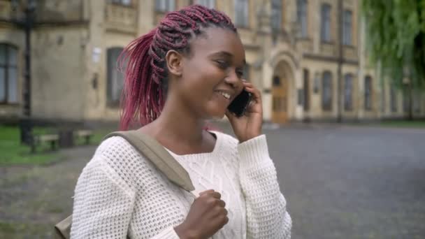 Young pretty african student talking on phone and laughing, standing with backpack in park near university, woman with pink dreadlocks