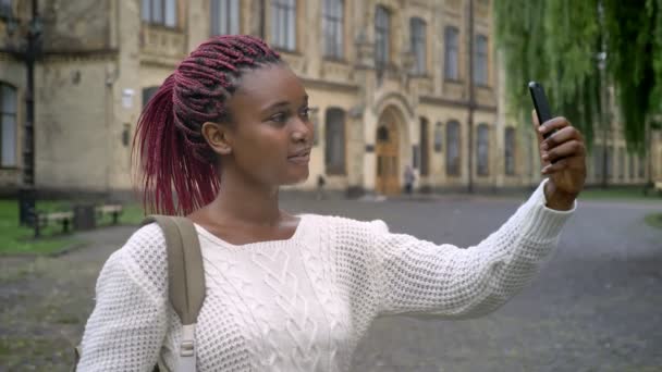 バックパックを押しながら公園に立っているピンク色のドレッドヘアを持つ女性、彼女の携帯電話で selfie を取って美しいアフリカの女子学生 — ストック動画