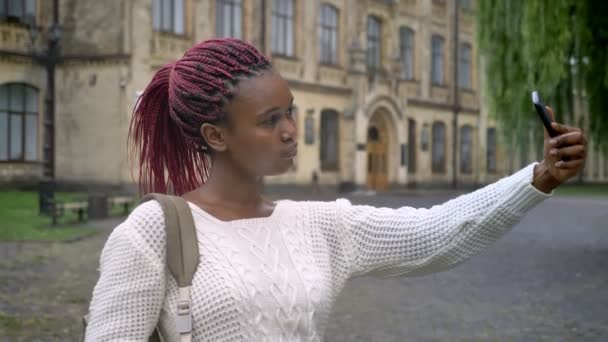 Joven encantadora mujer afroamericana tomando selfie y de pie con mochila en el parque cerca de la universidad — Vídeos de Stock