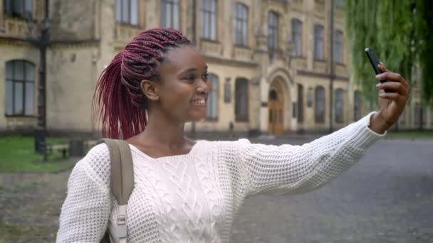 Beautiful african woman with pink dreadlocks taking selfie and standing with backpack in park near university — Stock Video