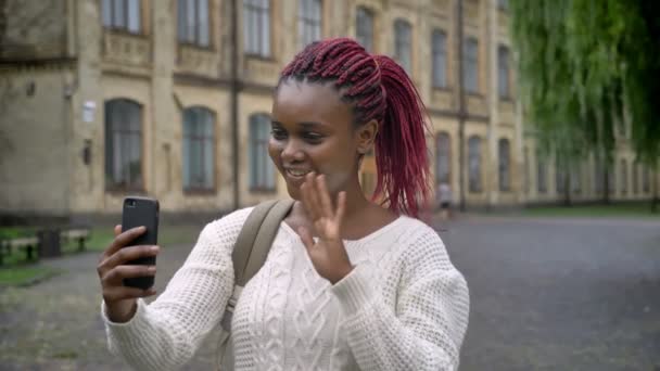 Hermosa joven africana estudiante teniendo video chat a través del teléfono y de pie en el parque cerca de la universidad, sonriendo — Vídeos de Stock