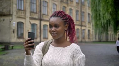 Smartphone üzerinden görüntülü sohbet sahip, konuşan ve gülen, sırt çantası ile neşeli kadın pembe dreadlocks ile genç Afrika öğrenci