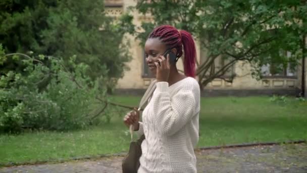 Joven hermosa estudiante saliendo de la universidad y hablando por teléfono, mujer con rastas rosadas caminando en el parque cerca de la universidad — Vídeo de stock