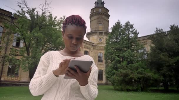 Unga afrikanska kvinnan med rosa dreadlocks i vit tröja maskinskrivning på tablett, sitter i parken nära university — Stockvideo