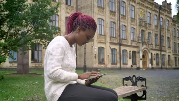 Młody african american Kobieta w białym swetrze przeglądania na tablecie i siedząc na ławce w parku, w pobliżu Uniwersytetu — Wideo stockowe