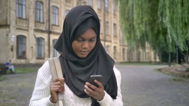 Sırt çantası tutan genç güzel Afrika Müslüman öğrenci telefonda sohbet ve üniversite, Parkı ayakta hijab içinde