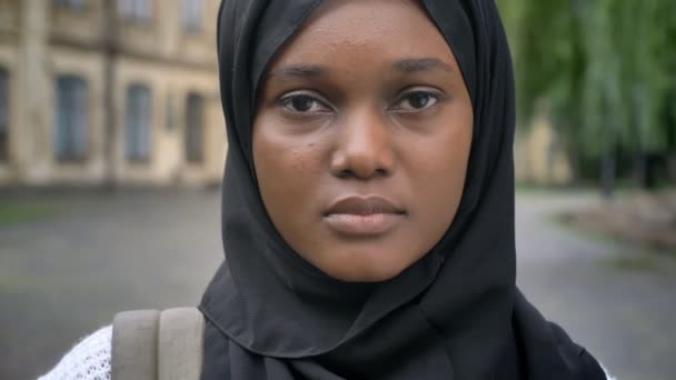 Traurige junge afrikanische Muslimin im Hidschab, die in die Kamera schaut und im Park in der Nähe des College steht, ernster und besorgter Ausdruck — Stockvideo