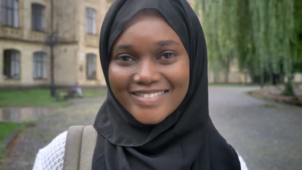 Charmante Afrikaanse jonge moslimvrouw in hijab op zoek en lachend op camera, permanent in park in de buurt van Universiteit, gelukkig en vrolijk — Stockvideo