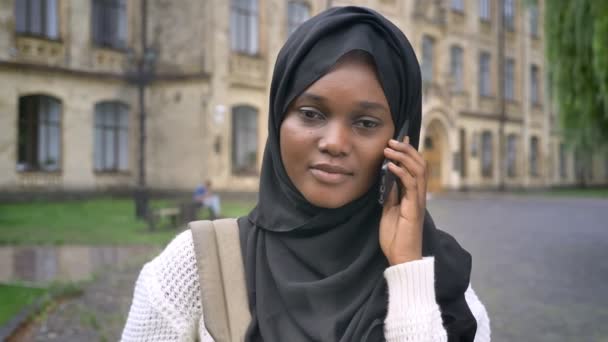 Portrait of african muslim student in hijab talking on cell phone and standing in park near university — Stock Video