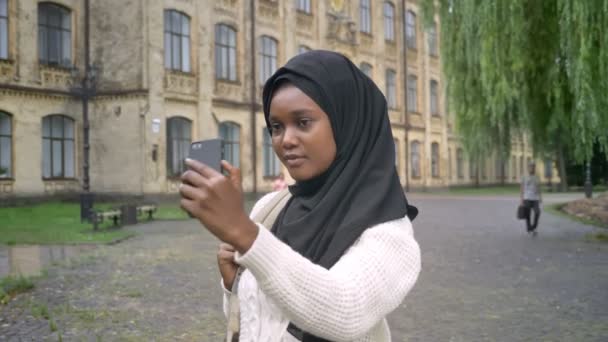 Joven mujer musulmana africana en hijab tomando selfie con teléfono y de pie en el parque cerca de la universidad, encantadora estudiante femenina con bolsa — Vídeos de Stock