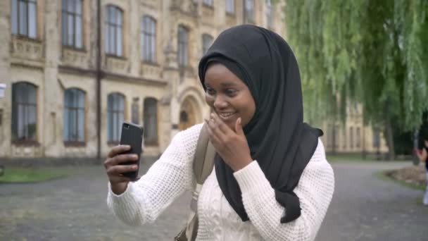 Jovem estudante muçulmano africano no hijab tendo vídeo chat através do telefone, de pé no parque perto da universidade, feliz e alegre — Vídeo de Stock