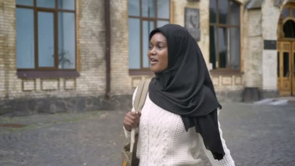 幸せで陽気な誰かに手を振って、大学近くの公園でウォーキング ヒジャーブの若いアフリカ系アメリカ人のイスラム教徒の女性 — ストック動画