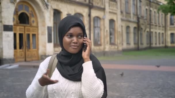 Encantadora joven musulmana africana en hijab hablando por teléfono celular y caminando en el parque cerca de la universidad, durante el día — Vídeo de stock