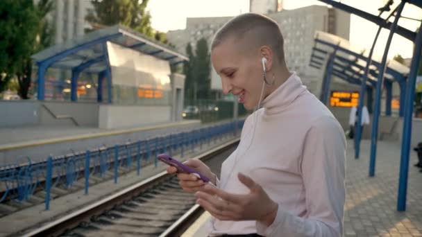 Young sad extraordinary bald girl is watching her phone and listening music on railway station in daytime in summer, urban concept — Stock Video