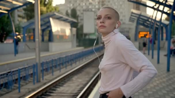 Triste mujer rubia calva caucásica está bailando en los auriculares mientras espera el tren en la plataforma durante el día — Vídeos de Stock